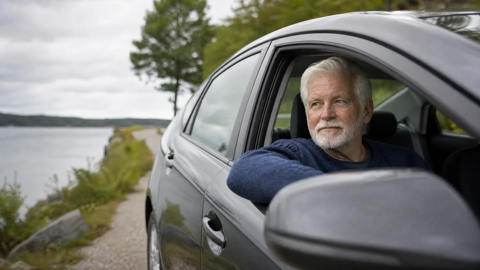 An older man with white hair and a beard, wearing a blue sweater, rests his arm on the open window of a dark car parked on a gravel road beside water and trees under an overcast sky.