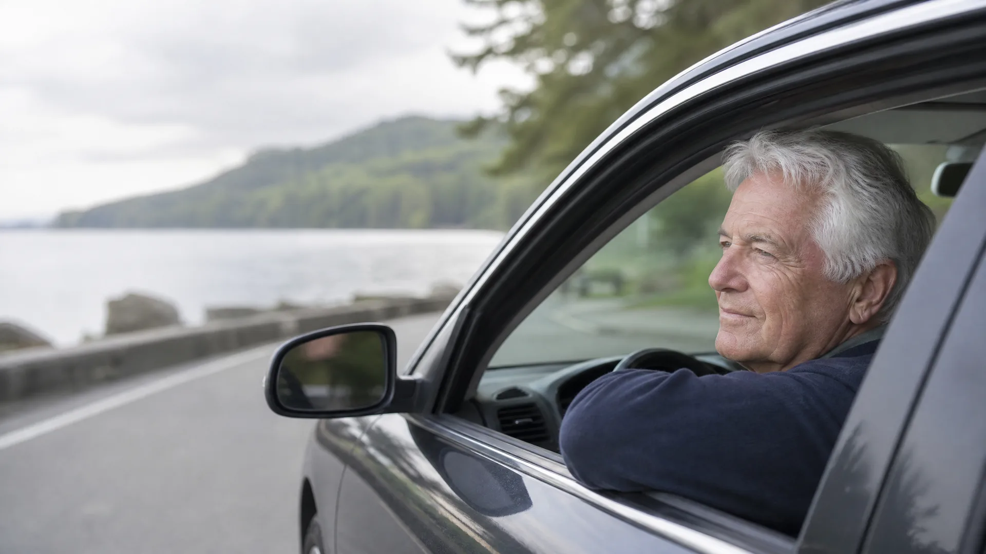 An older man with gray hair rests his arm on the open window of a car parked on a road beside water, with forested hills in the background.