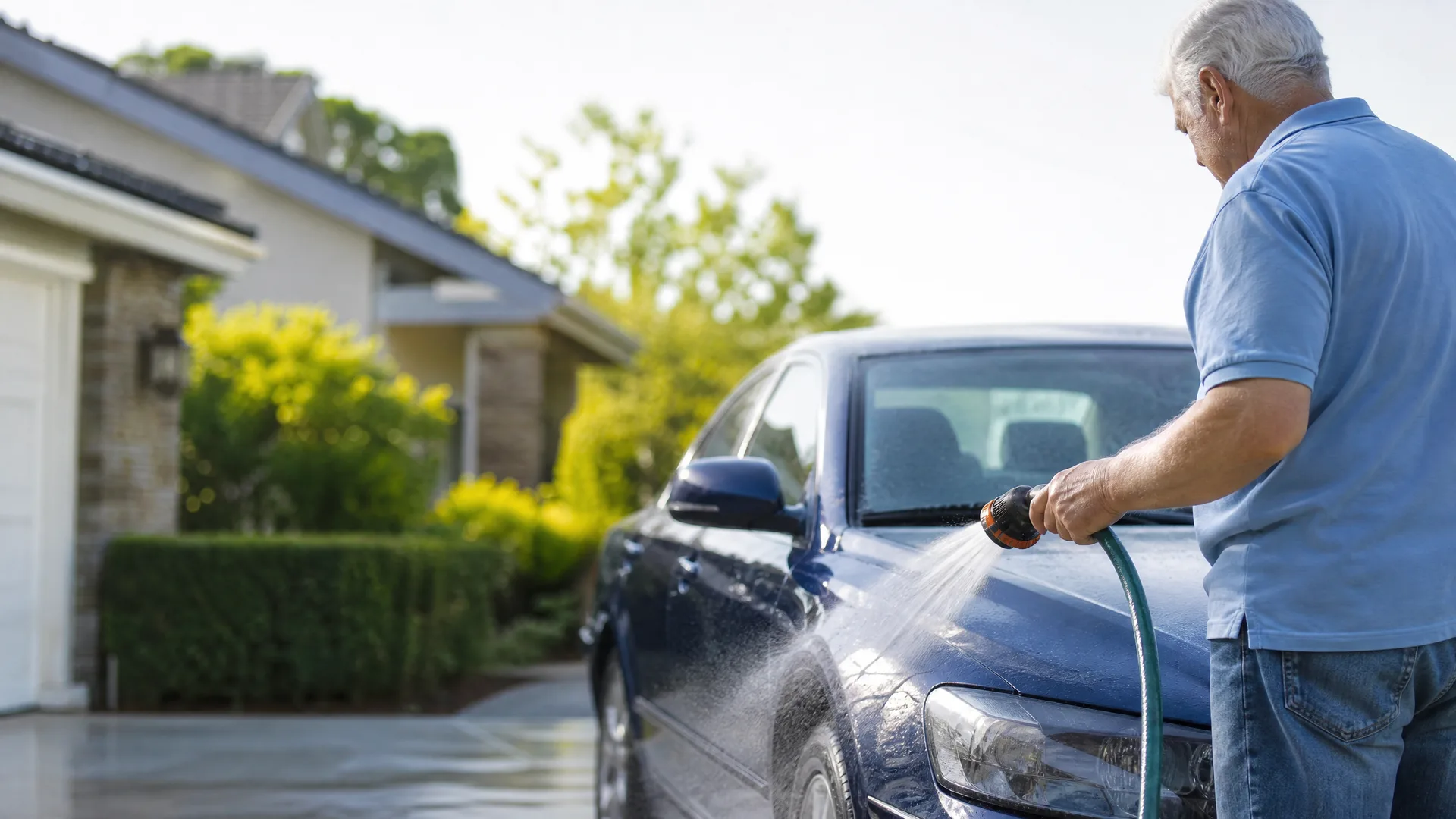 An older man in a light blue polo shirt sprays a dark blue sedan with a garden hose on a driveway in front of a single-story house with shrubs and trees.