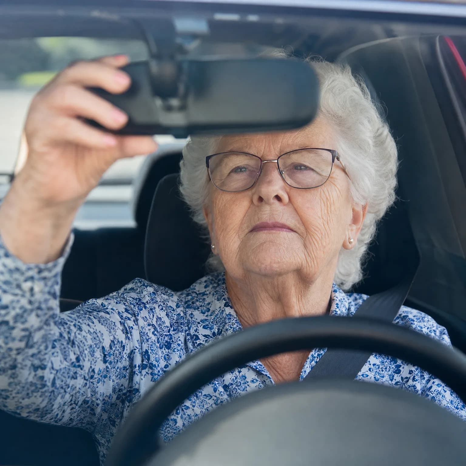 An elderly woman with white hair and glasses sits in a car's driver seat, reaching up with her left hand to adjust the rearview mirror, with the steering wheel blurred in the foreground.