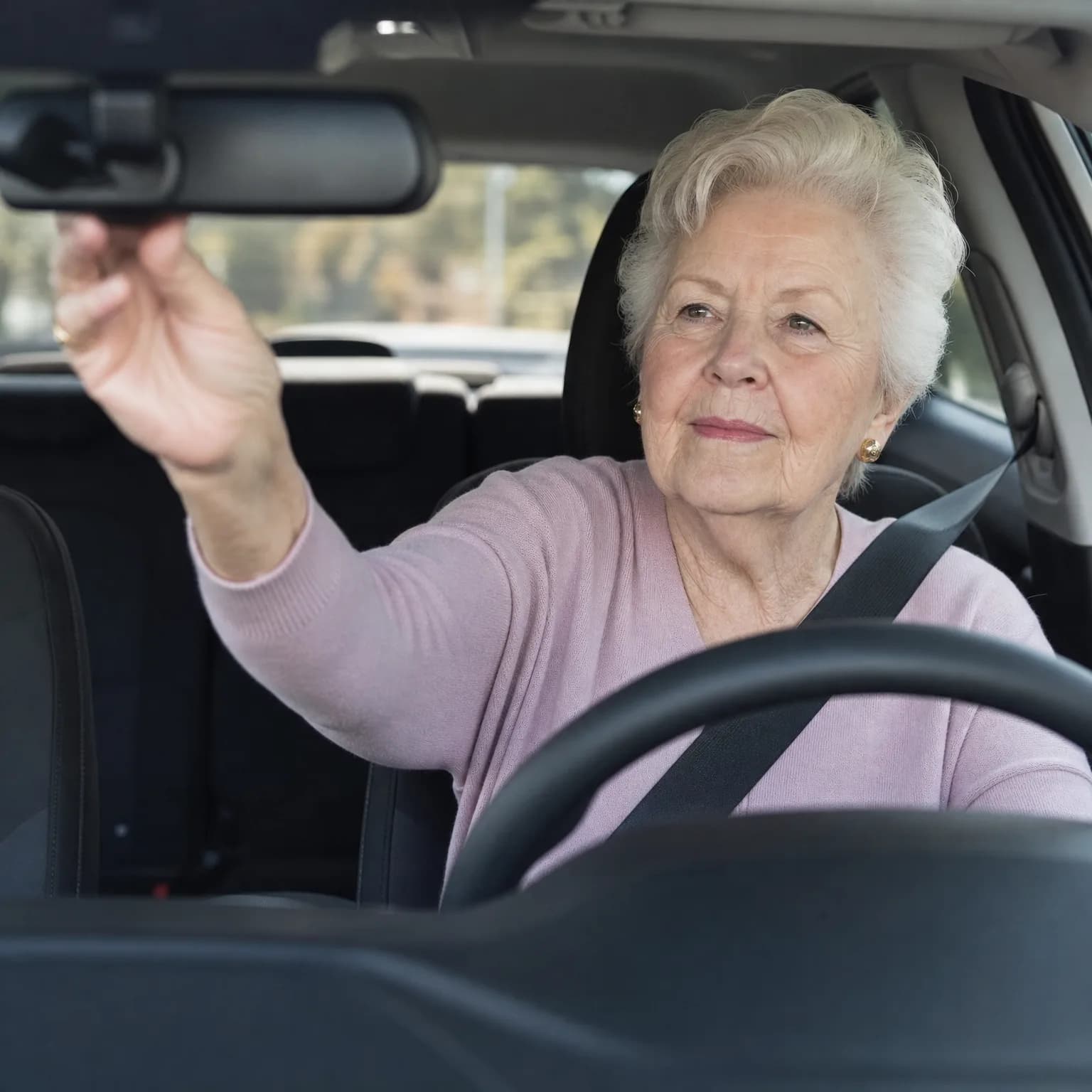 An older woman with short white hair in a pink sweater sits in a car's driver seat wearing a seatbelt and reaches up to adjust the rearview mirror.