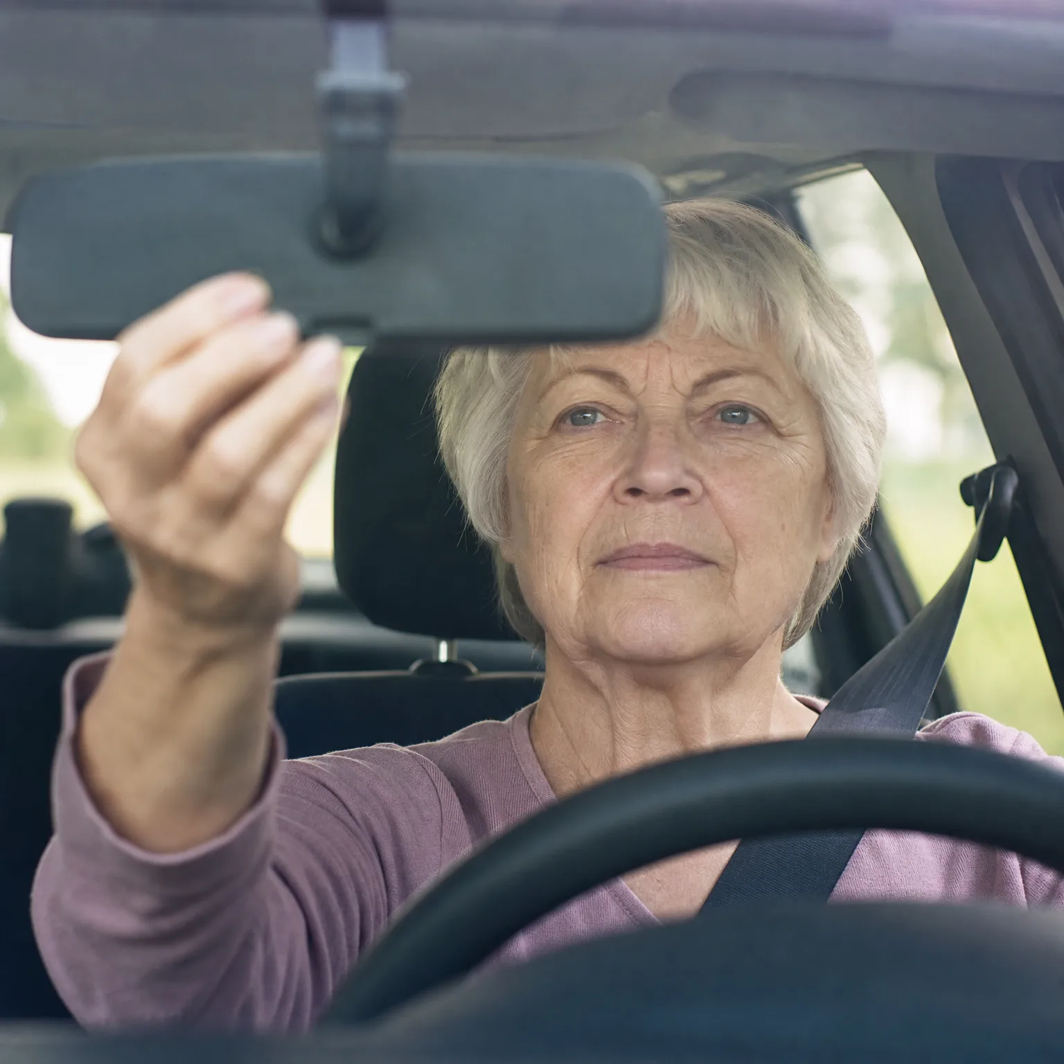 An older woman with short light hair, wearing a pink top and a seat belt, sits in the driver's seat of a car and reaches up to adjust the rearview mirror, seen through the windshield with green foliage outside the side window.
