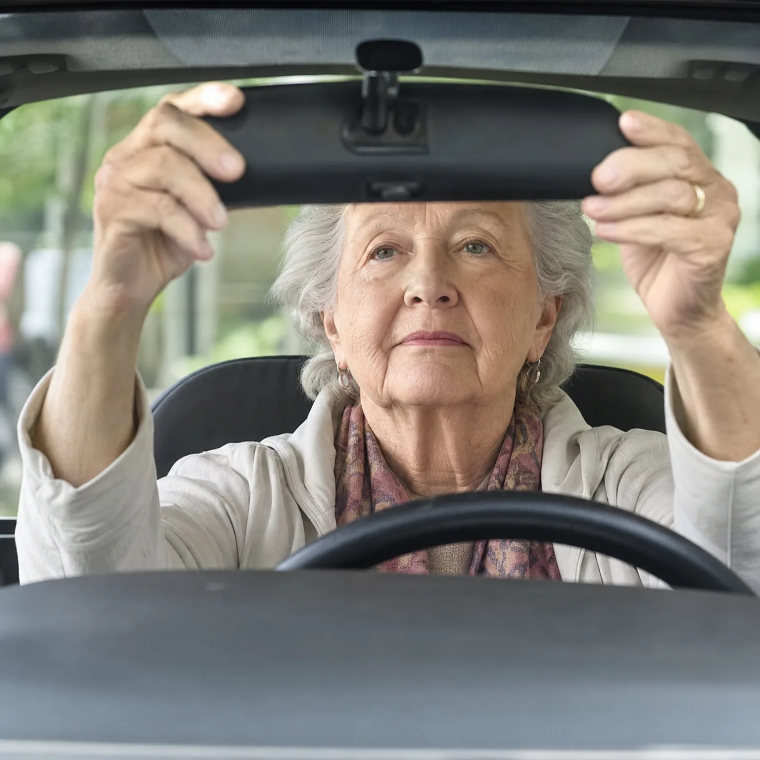 An older woman with gray hair, wearing a light jacket and patterned scarf, sits in a car's driver seat and adjusts the rearview mirror with both hands, seen through the windshield.