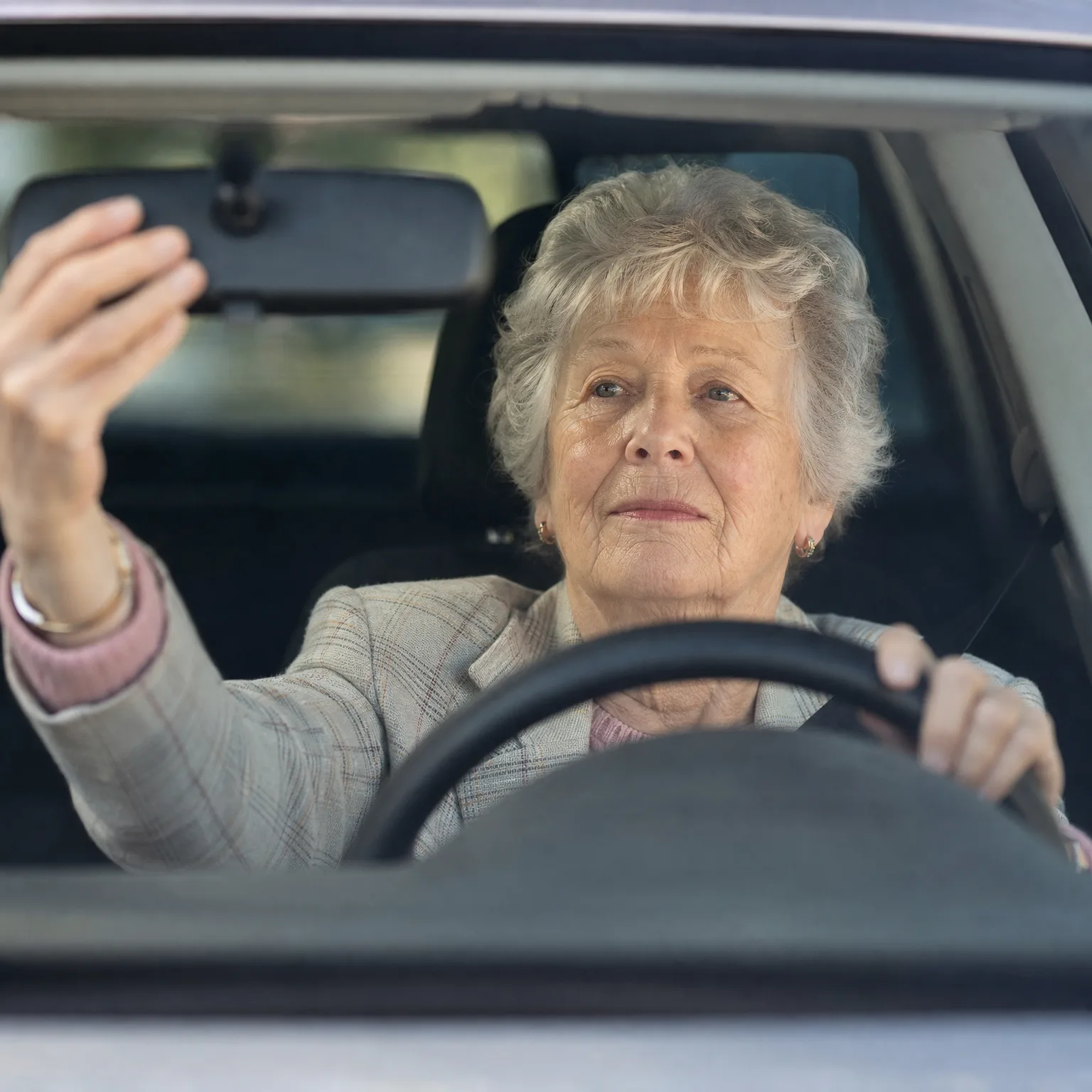 An older woman with short gray hair, seen through a windshield, holds the steering wheel with one hand while reaching up to adjust the rearview mirror with the other.
