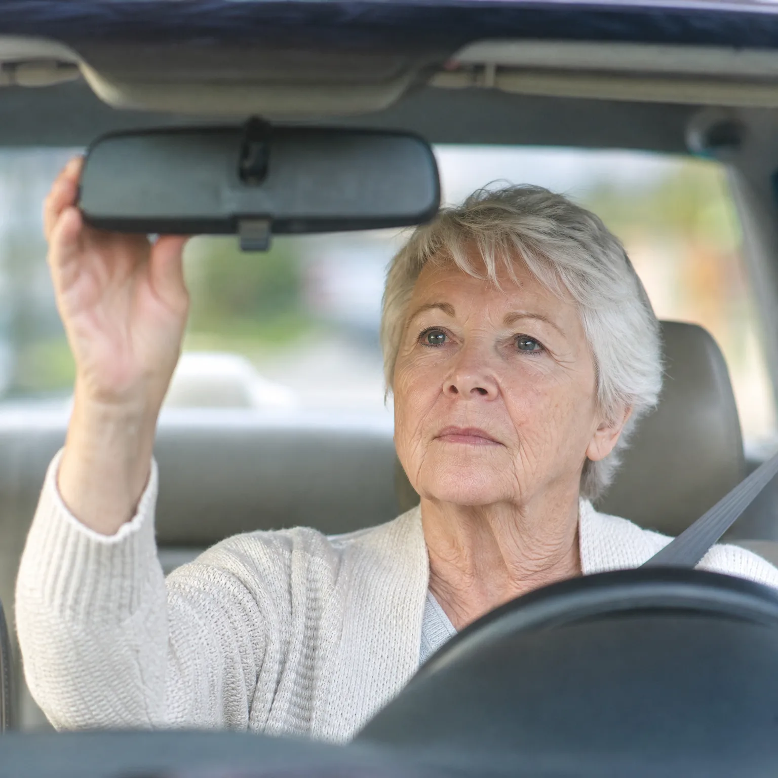 An older woman with short white hair, wearing a cream knit cardigan and seatbelt, reaches up to adjust the rearview mirror while seated in a car's driver seat.