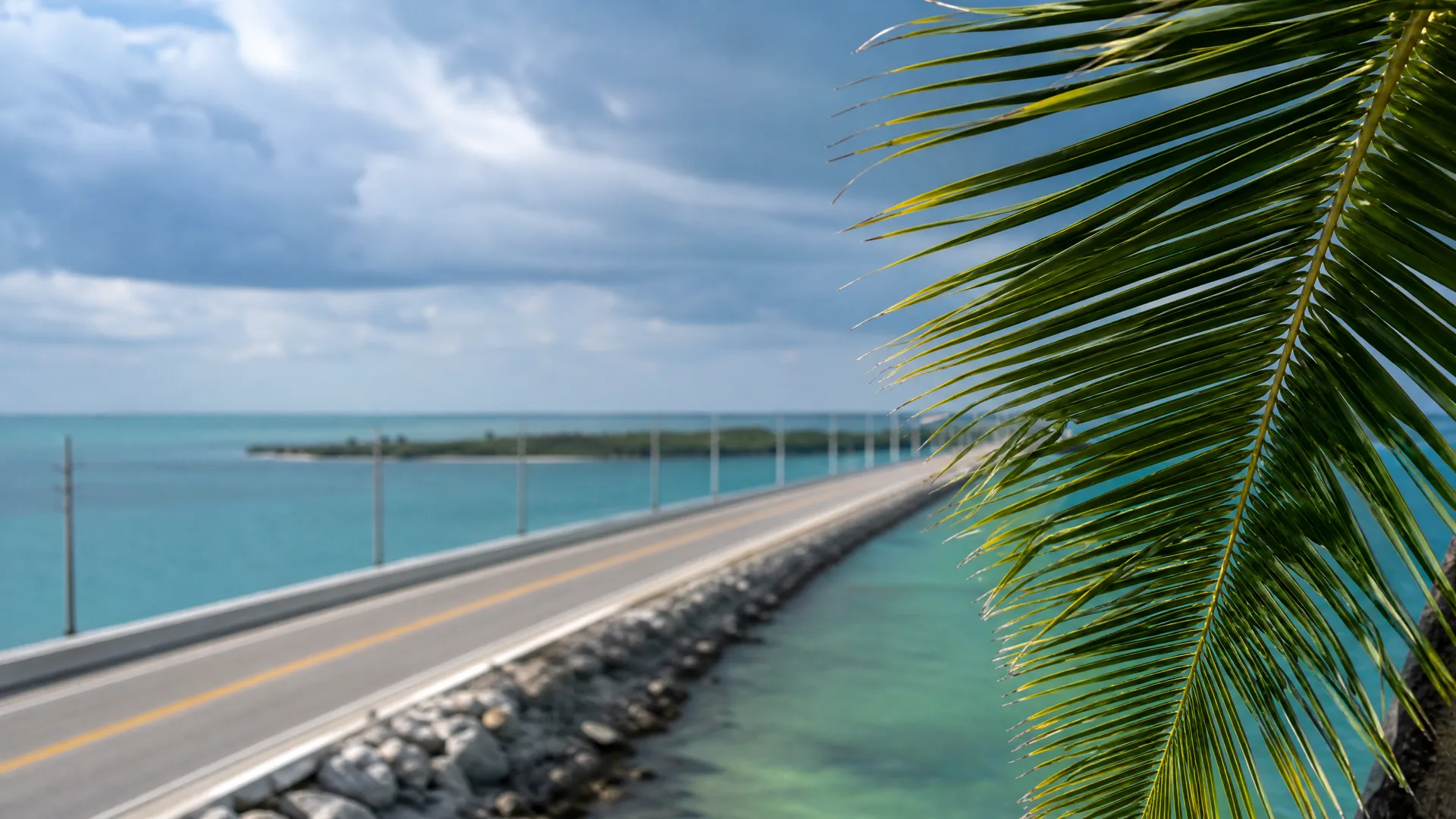 A two-lane highway on a causeway with a rock embankment runs over turquoise water toward a low island, with a palm frond in the foreground at right and cloudy sky above.