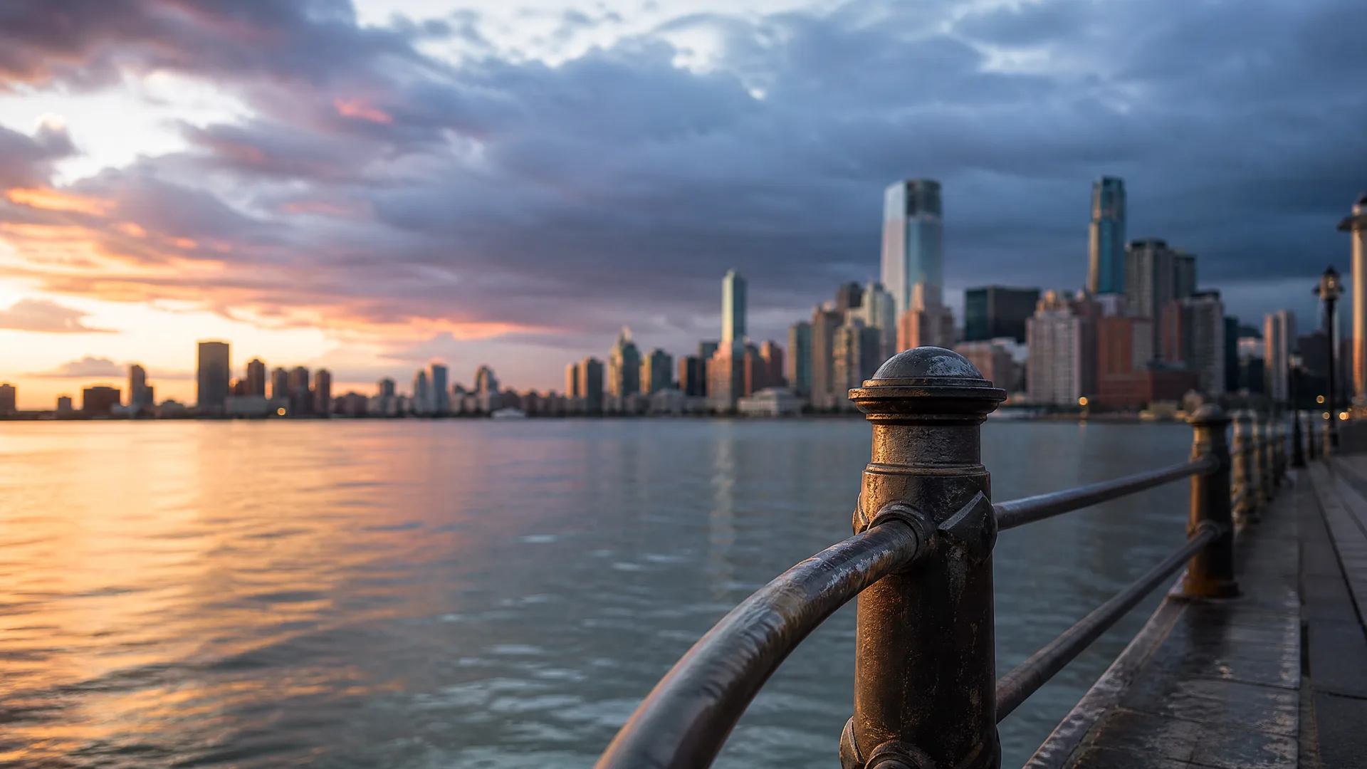 A weathered metal railing post in the foreground along a waterfront walkway, with a wide body of water and a distant city skyline under cloudy sky at sunset.
