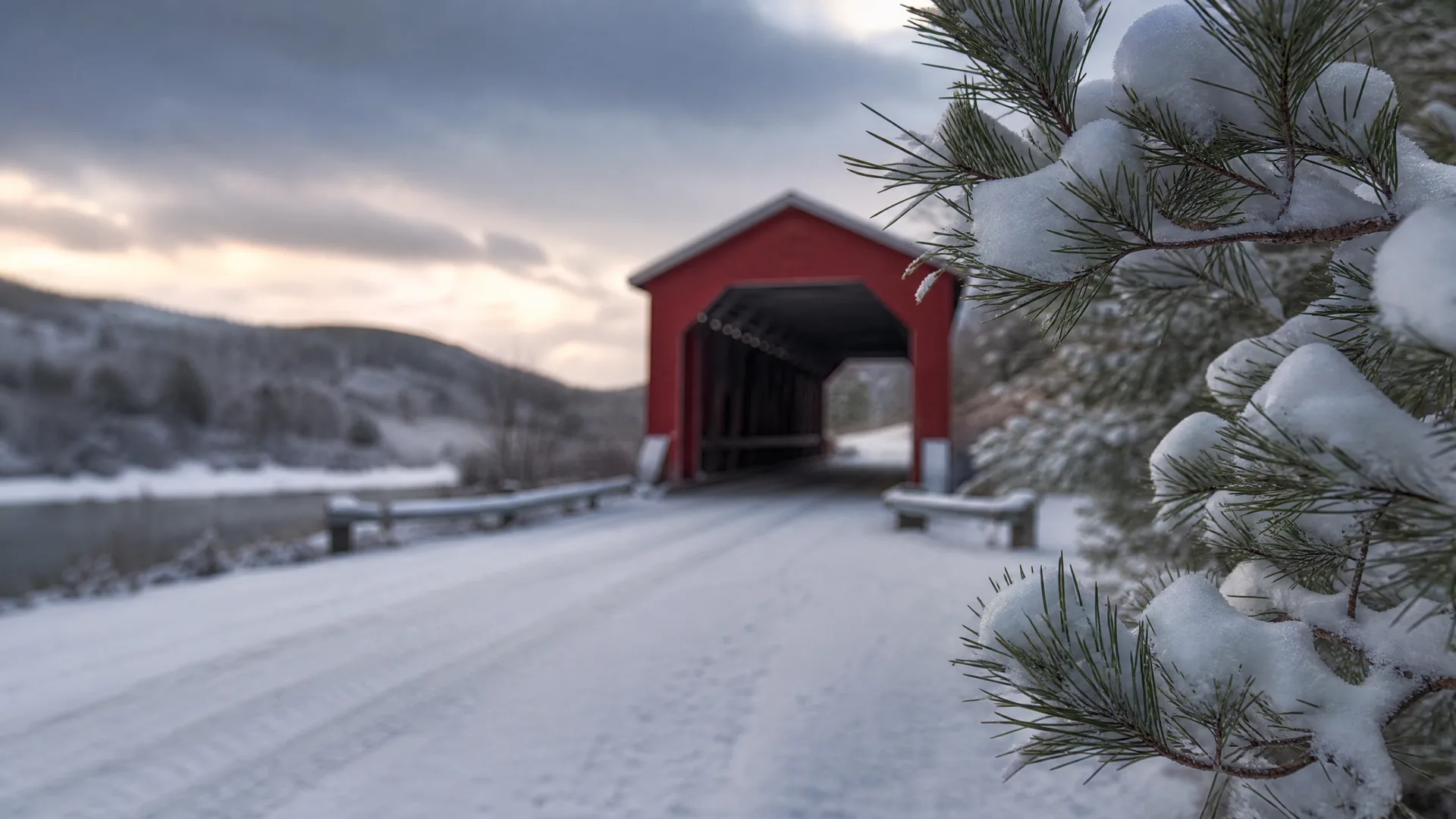 A red covered wooden bridge at the end of a snow-covered road, with a snow-laden pine branch in sharp focus in the foreground and low hills under an overcast sky behind.